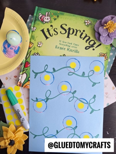 A spring-themed craft setup featuring a book titled Its Spring!, a yellow plate with a decorated egg, Plastic Egg Stamp Crafts, yellow stickers, felt flowers, and blue paper with a floral drawing. Text: @GLUEDTOMYCRAFTS.