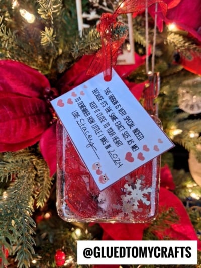 A handmade Christmas ornament with a snowflake and red background hangs on the tree. Attached is a note, hearts, and a height poem about reading, signed “Love, Stacey, Xmas 2024.” Large red poinsettias decorate the tree.