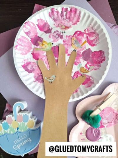 A craft project featuring a paper plate with pink paint and bird stickers, a cutout brown paper hand, and spring-themed decorations captures the charm of cherry blossom tree crafts, with a Hello Spring notepad and art supplies.