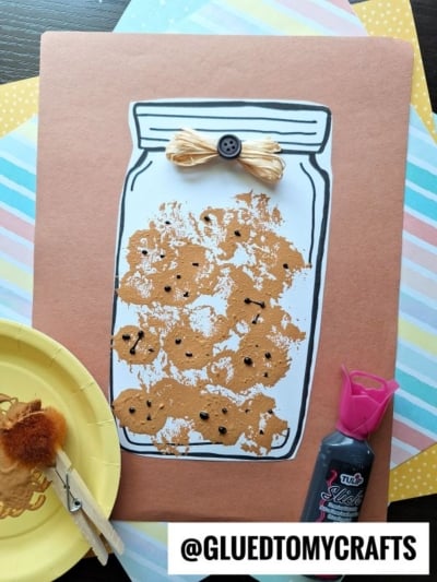 A cookie jar craft featuring a mason jar filled with brown pom pom stamped cookie prints, adorned with black dots and a twine bow. Nearby are a paint plate, clothespin brush, and black paint bottle. @GLUEDTOMYCRAFTS is at the bottom.