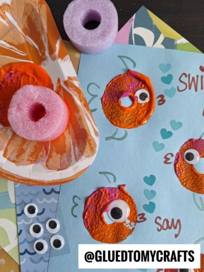 A blue paper with orange, fish-like prints made using a painted pool noodle, each with a googly eye, surrounded by cut-out shapes and the words swim, say, and hearts. @GLUETOMYCRAFTS watermark at the bottom.