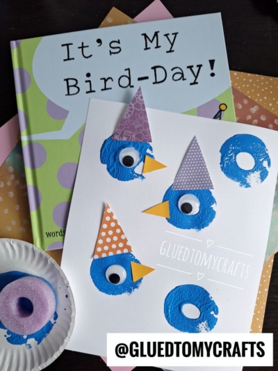 A children’s book titled “It’s My Bird-Day!” is next to a craft with blue painted birds made from donut-shaped stamps, googly eyes, and patterned paper hats. @GLUEDTOMYCRAFTS is written on the craft.