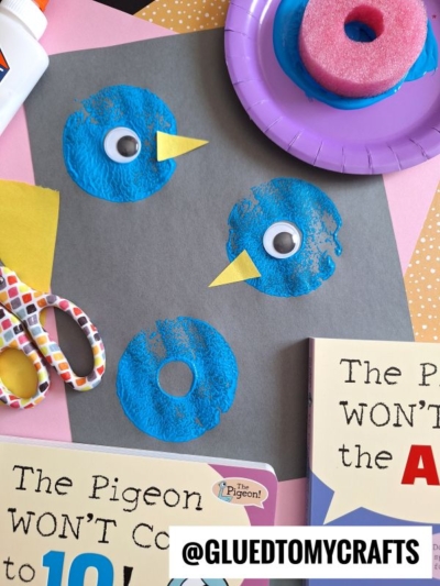 Children’s craft project featuring blue sponge-painted circles with googly eyes and yellow paper beaks on black paper, surrounded by craft supplies, scissors, glue, and The Pigeon books by Mo Willems.