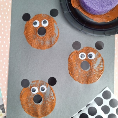 Three bear faces are stamped on gray paper using brown paint circles, black ears, and googly eyes. Craft supplies, including foam and stickers, are visible around the sheet. The image is watermarked @GLUETOMYCRAFTS.