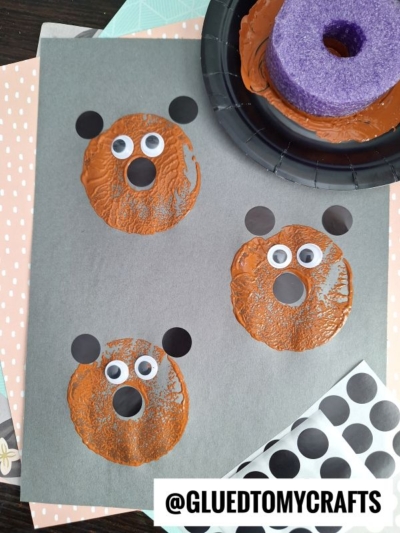 Three bear faces are stamped on gray paper using brown paint circles, black ears, and googly eyes. Craft supplies, including foam and stickers, are visible around the sheet. The image is watermarked @GLUETOMYCRAFTS.