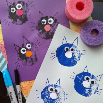 Two sheets of paper show cat faces stamped with paint and a pool noodle. Each cat has googly eyes, drawn whiskers and ears, and a pom-pom nose. Markers and a pool noodle are nearby. @GLUEDTOMYCRAFTS is labeled at bottom.