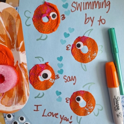 A blue sheet with four orange fish made from paint and googly eyes, hearts drawn around them, and the handwritten message: “Swimming by to say I love you!” Markers and craft supplies are nearby.