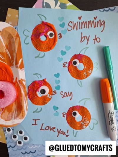 A blue sheet with four orange fish made from paint and googly eyes, hearts drawn around them, and the handwritten message: “Swimming by to say I love you!” Markers and craft supplies are nearby.