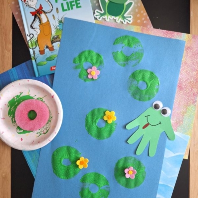 A children’s pond life craft features lily pads and flowers made from green paint prints on blue paper, with a frog cut from green paper. A Dr. Seuss book, a paper frog, and crafting materials are nearby.