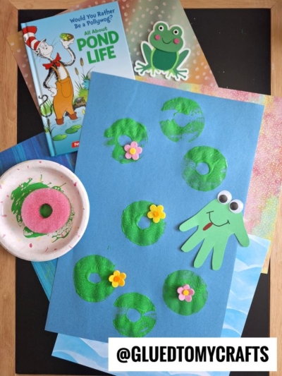 A children’s pond life craft features lily pads and flowers made from green paint prints on blue paper, with a frog cut from green paper. A Dr. Seuss book, a paper frog, and crafting materials are nearby.