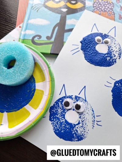 A blue cat craft made with a pool noodle stamp, blue paint, googly eyes, and paper is shown alongside a book and crafting supplies. The Instagram handle @GLUEDTOMYCRAFTS is visible at the bottom.