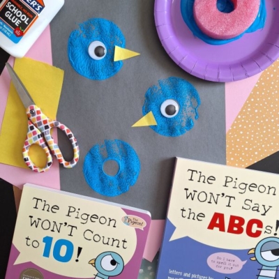 A craft scene features blue pigeon art made from painted sponges on black paper, googly eyes, and yellow beaks. Surrounding items include glue, scissors, colored paper, two Pigeon books by Mo Willems, and a plate with a paint-covered sponge.
