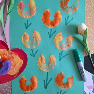 A green sheet of paper shows orange and yellow painted flower shapes stamped with a foam donut, green stems drawn with marker, and artificial tulips nearby. Supplies and a @gluedtomycrafts label are also visible.