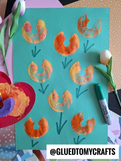 A green sheet of paper shows orange and yellow painted flower shapes stamped with a foam donut, green stems drawn with marker, and artificial tulips nearby. Supplies and a @gluedtomycrafts label are also visible.