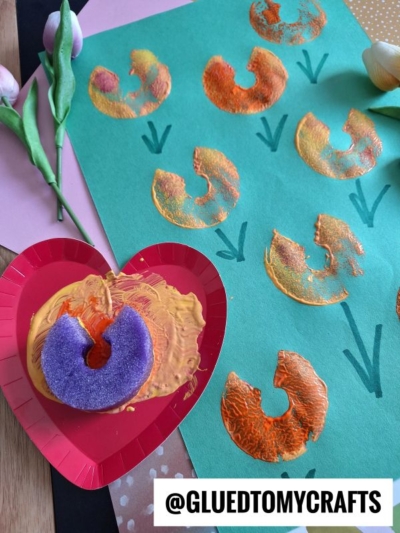 A green paper with orange and yellow painted tulip shapes stamped in rows, created using a purple pool noodle dipped in paint. Green stems are drawn under each flower. A heart-shaped tray holds paint and the noodle.