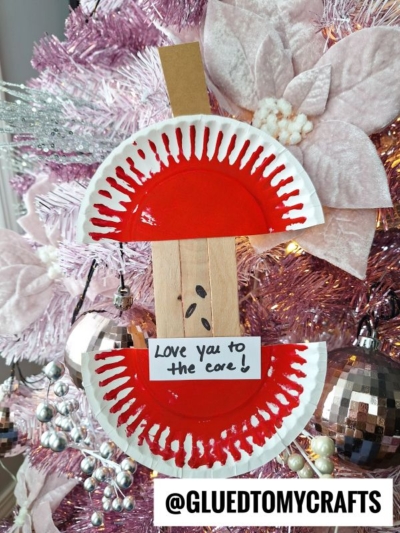 A red and white paper plate mother’s day apple core craft with a wooden stick for a stem hangs on a pink Christmas tree. A note on the craft reads, Love you to the core! @GLUEDTOMYCRAFTS is displayed at the bottom.