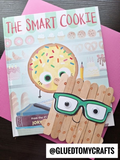 A children’s book titled The Smart Cookie is shown with a googly-eyed, glasses-wearing cookie craft made from popsicle sticks—perfect for back to school story time. The items rest on pink polka dot paper. @GLUEDTOMYCRAFTS is written at the bottom.