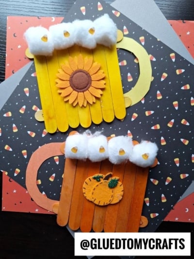 Two craft mugs made from colored popsicle sticks, topped with cotton to resemble foam, decorated with a felt sunflower and pumpkin, are perfect FALL CRAFTS FOR KIDS and placed on Halloween-themed paper with candy corn patterns. @GLUEDTOMYCRAFTS is written at the bottom.