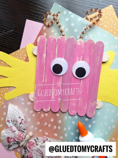 A pink popsicle stick insect craft with large googly eyes sits on colorful polka-dot paper, yellow hand-shaped cutouts, a paper butterfly, pipe cleaners, and a bottle of glue—perfect for exploring fun INSECT CRAFT IDEAS! @GLUEDTOMYCRAFTS is written at the bottom.