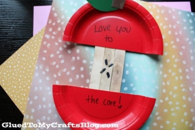 A craft made from red paper plates forms a sliced apple, with “Love you to the core!” written inside. The background includes colorful, polka-dot papers. GluedToMyCraftsBlog.com appears at the bottom.
