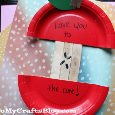 A craft made from red paper plates forms a sliced apple, with “Love you to the core!” written inside. The background includes colorful, polka-dot papers. GluedToMyCraftsBlog.com appears at the bottom.