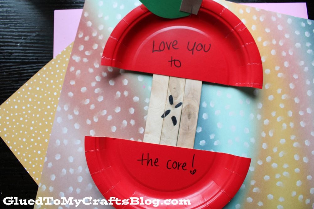 A craft made from red paper plates forms a sliced apple, with “Love you to the core!” written inside. The background includes colorful, polka-dot papers. GluedToMyCraftsBlog.com appears at the bottom.