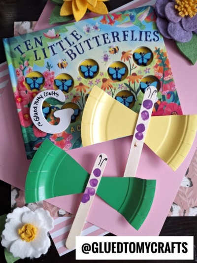 Colorful children’s book Ten Little Butterflies sits beside two popsicle stick butterfly crafts made from paper plates, displayed on pink paper with felt flower decorations. @GLUEDTOMYCRAFTS is written at the bottom.