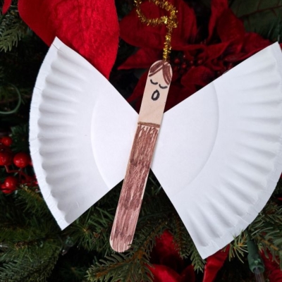 A handmade angel ornament with wings made from a cut paper plate, a craft stick body colored brown, a drawn face, and a gold pipe cleaner halo, hanging on a Christmas tree.