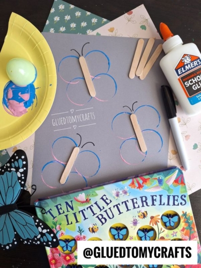 A butterfly craft activity with colored paper, popsicle sticks, glue, marker, and a butterfly book arranged on a table. The centerpiece features gray paper with butterfly outlines and popsicle sticks, alongside fun Plastic Egg Stamp Crafts.