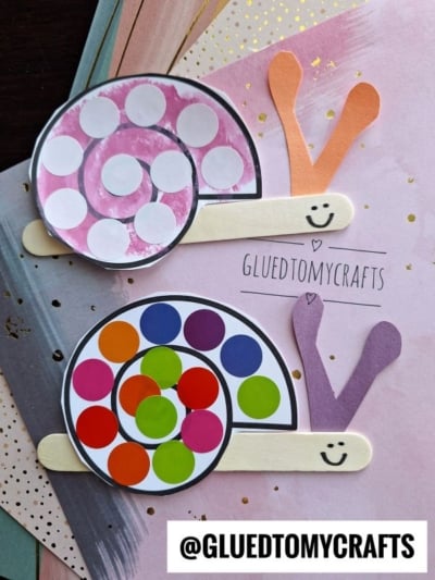 Two paper snails made from colored circles, craft sticks, and construction paper sit on a pastel background. Each snail features a smiling face and colorful spiral shells. The handle @GLUEDTOMYCRAFTS is visible on the image.