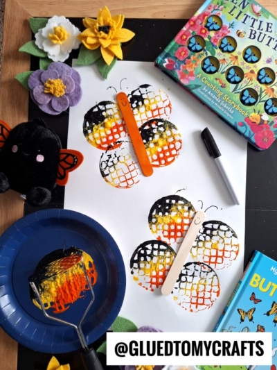 A kids’ craft scene with a paper showing painted bees made from potato masher stamped craft and popsicle stick bodies, flowers, plush bee, paint roller, books about butterflies, and an @GLUEDTOMYCRAFTS tag.