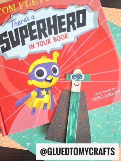 A children’s book titled “There’s a Superhero in Your Book” lies on a green patterned surface. In front is a craft superhero figure—one of many fun summer craft ideas—made from a popsicle stick, green mask, and paper cape.