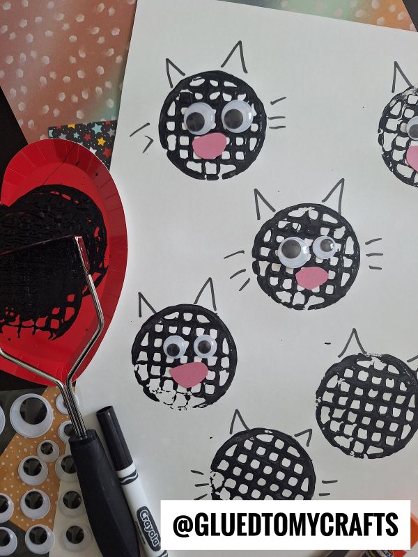 Stamped black cat faces with googly eyes, pink noses, and whiskers on white paper, created using a black flyswatter, a red plate, a marker, and craft supplies. @GLUEDTOMYCRAFTS is printed at the bottom.