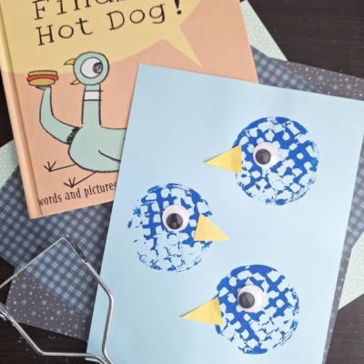 A children’s book titled The Pigeon Finds a Hot Dog! sits next to a blue paper with three potato masher stamped pigeons. The birds have googly eyes and yellow beaks, with craft tools and patterned papers in the background.