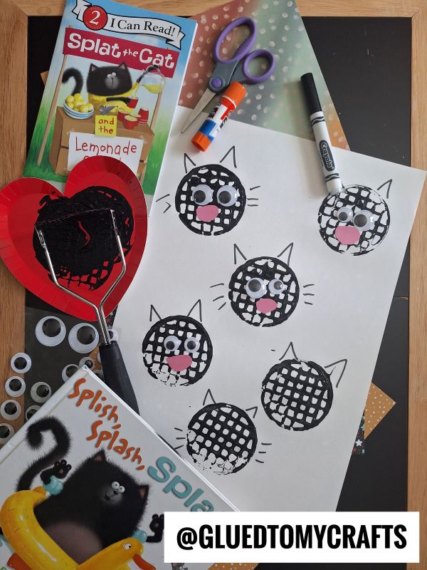 A kids’ craft setup with Splat the Cat books, googly eyes, scissors, a heart-shaped spatula, and a sheet with black cat faces made from paint and paper cutouts. @GLUEDTOMYCRAFTS appears at the bottom.