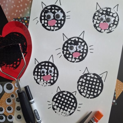 A sheet of paper with black cat faces made from paint and a potato masher, decorated with googly eyes and pink paper noses. Craft supplies including a marker, glue stick, and googly eyes are nearby.