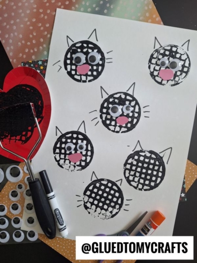 A sheet of paper with black cat faces made from paint and a potato masher, decorated with googly eyes and pink paper noses. Craft supplies including a marker, glue stick, and googly eyes are nearby.