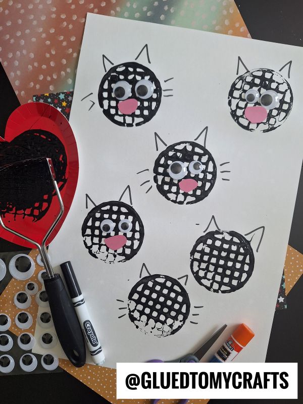 A sheet of paper with black cat faces made from paint and a potato masher, decorated with googly eyes and pink paper noses. Craft supplies including a marker, glue stick, and googly eyes are nearby.