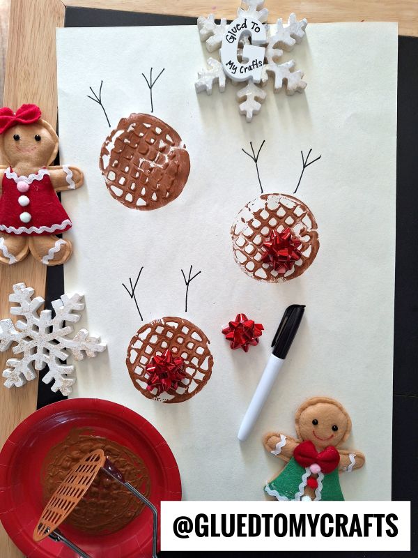 A piece of paper with brown paint stamped circles resembling reindeer heads, antlers drawn with black marker, red bows as noses, surrounded by gingerbread and snowflake ornaments, and painting supplies.