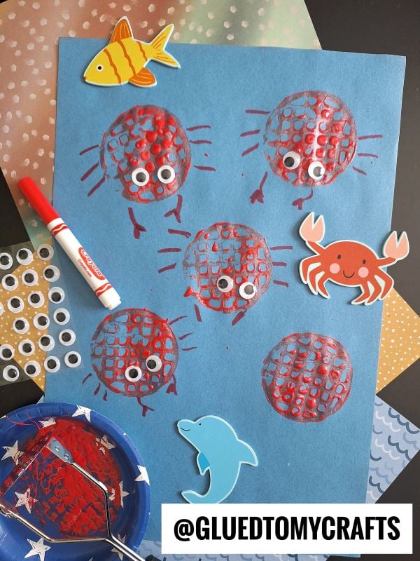 A blue paper displays five red crab prints made with mesh circles, each with googly eyes and drawn legs. Surrounding the paper are ocean-themed stickers and craft supplies, including a marker and paint.