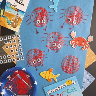 Blue paper with red crab prints made using a round stamp, googly eyes glued on each crab. Surrounded by Pete the Cat books, craft supplies, paper fish, crab, dolphin, and a paint roller on a black surface.