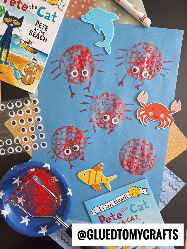 Blue paper with red crab prints made using a round stamp, googly eyes glued on each crab. Surrounded by Pete the Cat books, craft supplies, paper fish, crab, dolphin, and a paint roller on a black surface.