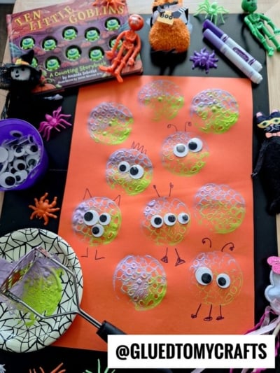 A Halloween craft setup featuring bubble wrap paint prints and potato masher stamped monsters on orange paper, decorated with googly eyes and marker details. Craft supplies, toy bugs, and the book Ten Little Goblins surround the artwork.