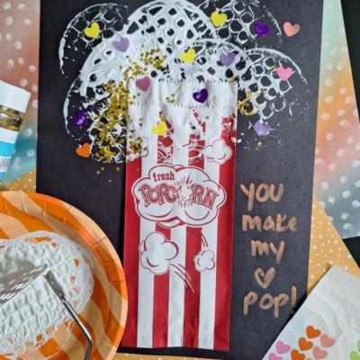 A potato masher stamped craft featuring a popcorn bag on black paper with white paint resembling popcorn, colorful paper hearts, and gold glitter. The words You make my heart pop! are written in gold. Craft supplies are nearby.