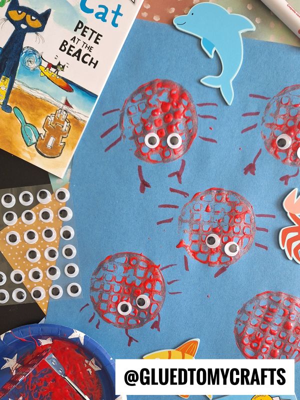 A blue paper with red crab prints made from mesh, each with googly eyes and drawn legs, surrounded by craft supplies, a dolphin cutout, and a Pete the Cat book.