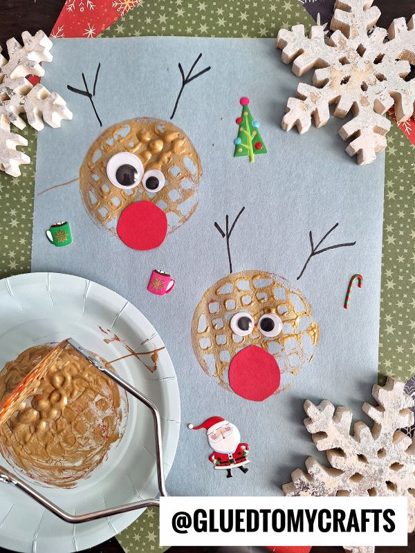 Two reindeer faces made from textured gold paint circles on blue paper, with googly eyes, red paper noses, and antlers drawn in black. Surrounded by festive stickers, snowflakes, and a craft stamp.