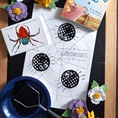 A children’s craft setup with a Charlotte’s Web book, a spider illustration, a spiderweb drawing with three black spotted spiders, purple and yellow felt flowers, a plate with black paint and roller, plus a potato masher stamped craft on a wooden tray.
