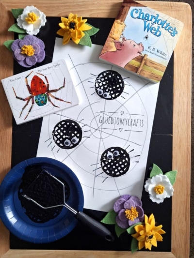 A children’s craft setup with a Charlotte’s Web book, a spider illustration, a spiderweb drawing with three black spotted spiders, purple and yellow felt flowers, a plate with black paint and roller, plus a potato masher stamped craft on a wooden tray.