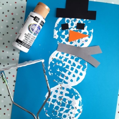 A snowman made with white paint and a potato masher on blue paper, decorated with black hat, orange nose, gray scarf cutouts, and surrounded by craft supplies including paint and a masher. @GLUEDTOMYCRAFTS is labeled at the bottom.