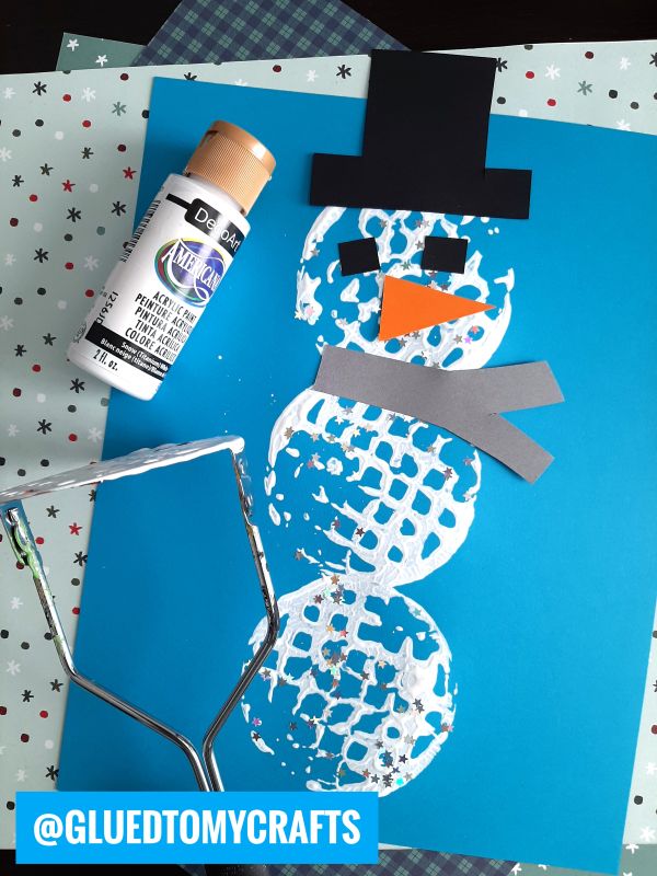 A snowman made with white paint and a potato masher on blue paper, decorated with black hat, orange nose, gray scarf cutouts, and surrounded by craft supplies including paint and a masher. @GLUEDTOMYCRAFTS is labeled at the bottom.